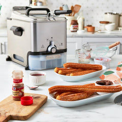 A compact deep fryer on a kitchen counter, surrounded by dishes of freshly made churros, seasonings, and baking tools