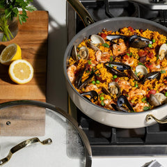 A jumbo cooker on a gas stove filled with seafood paella. Mussels, shrimp, and peas are visible. Sliced lemons sit on a wooden cutting board nearby.