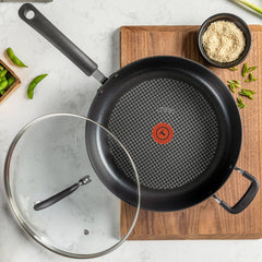 Non-stick frying pan on wooden cutting board beside ingredients, glass lid removed.