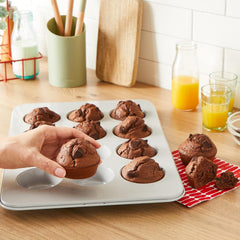 Hand removing chocolate muffins from muffin tray; orange juice on kitchen counter.