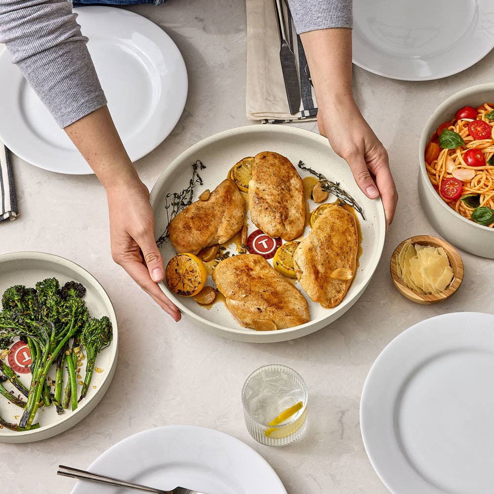 Hands hold a beige pan of roasted chicken and lemons surrounded by dishes and empty plates
