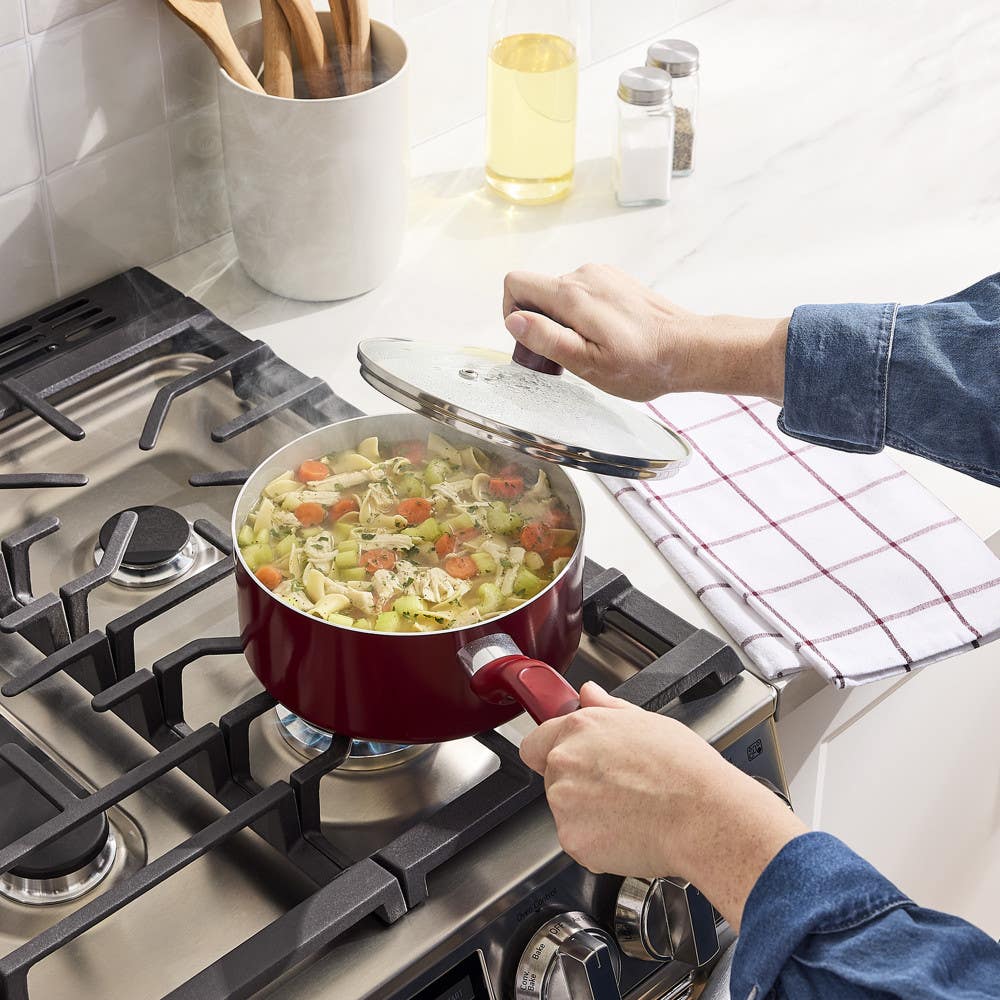 Hand lifting glass lid from red stockpot filled with steaming vegetable pasta on gas stove
