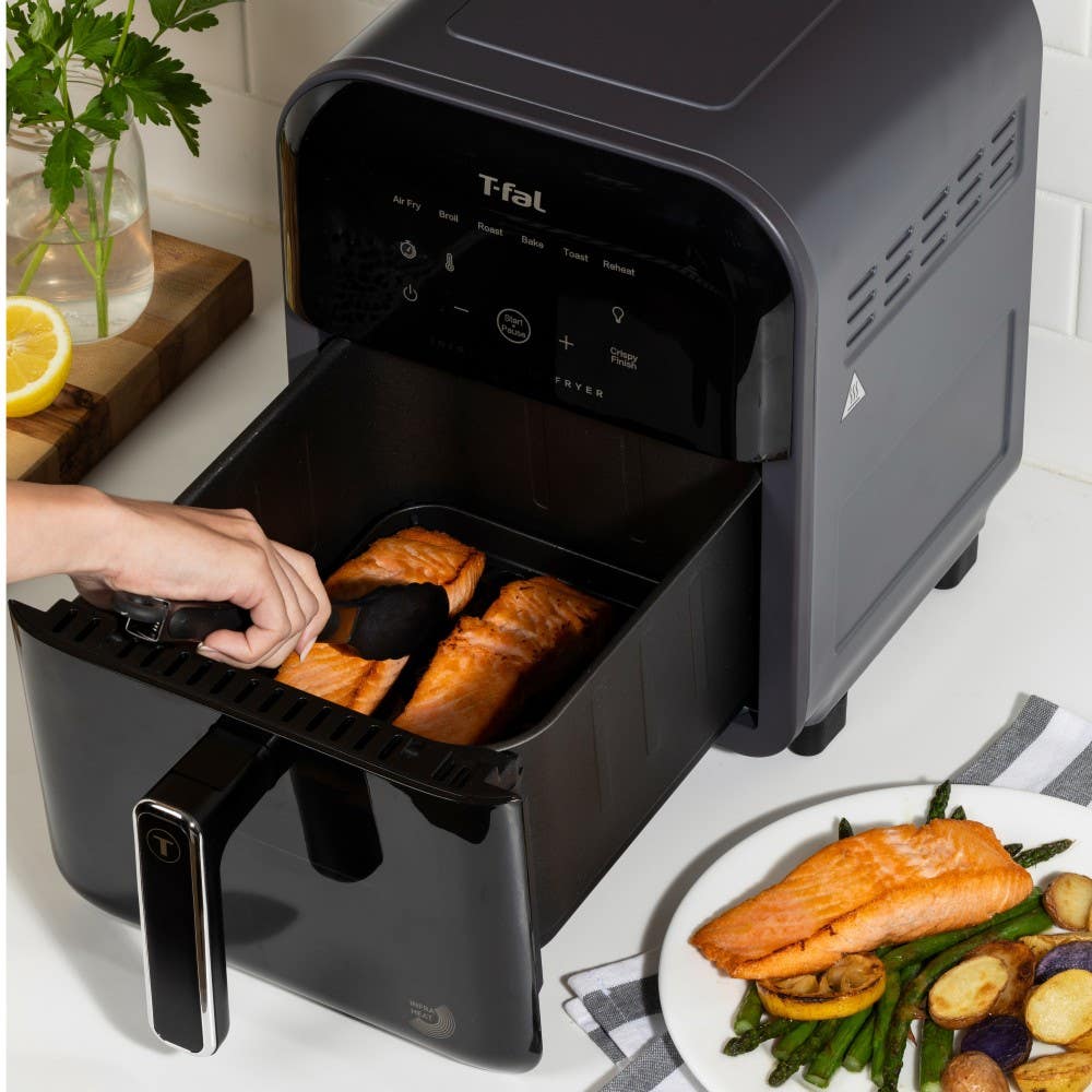 A person uses a spatula to handle salmon fillets in a T-fal infrared air fryer. Beside it, a nicely plated dish of cooked salmon accompanied by roasted vegetables is visible.