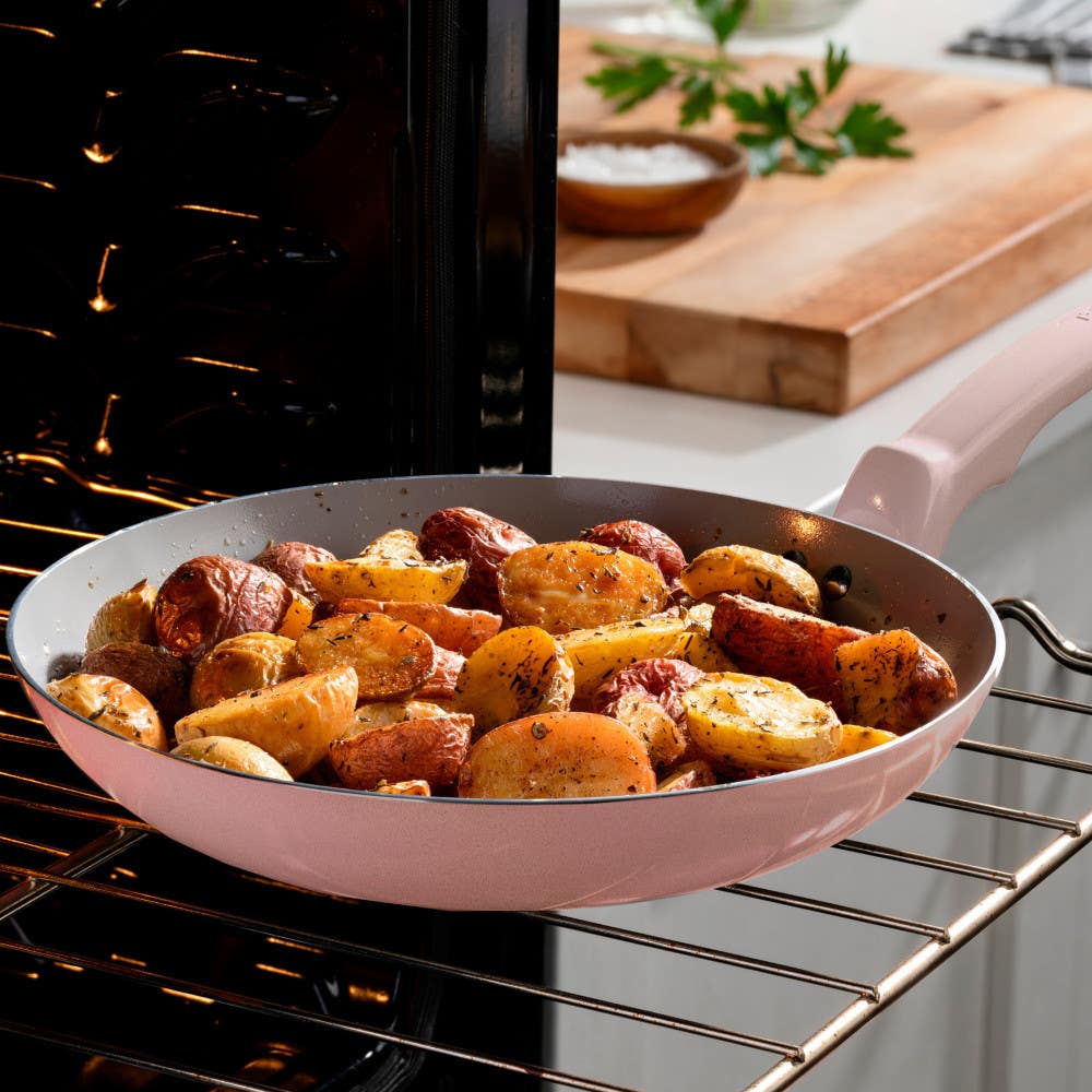 A pink frypan with roasted potatoes in an oven, a cutting board in the background