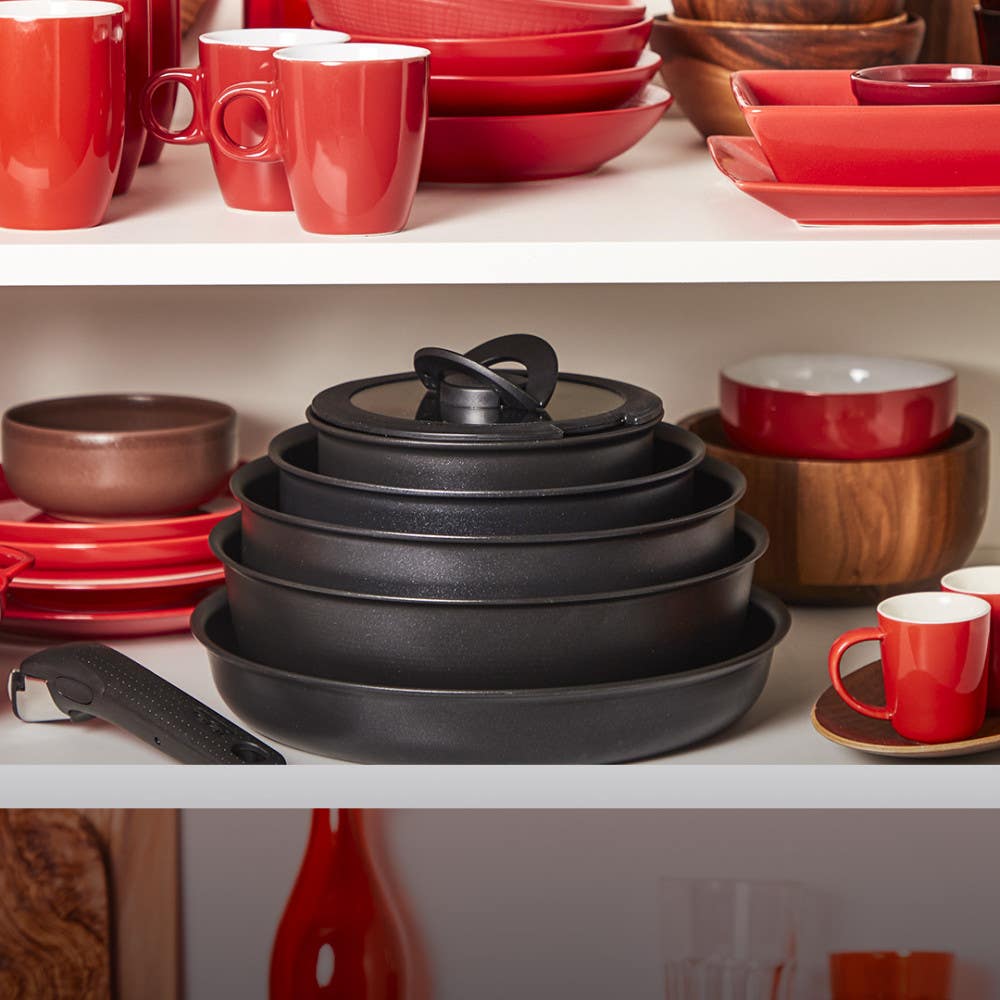 Stacked black cookware among red and white dishware in a cabinet and a detachable handle