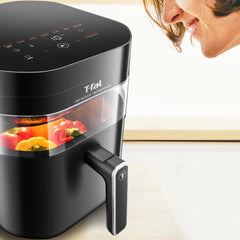 A woman looks at an air fryer with a built-in window, showing colorful vegetables cooking inside, promoting the convenient window feature for easy monitoring.