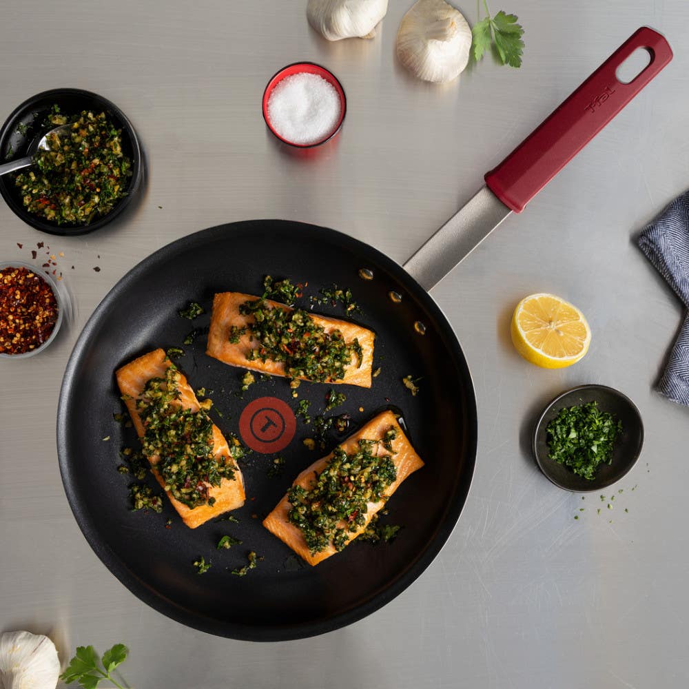 A red-handled pan with three seasoned salmon fillets, surrounded by cooking ingredients