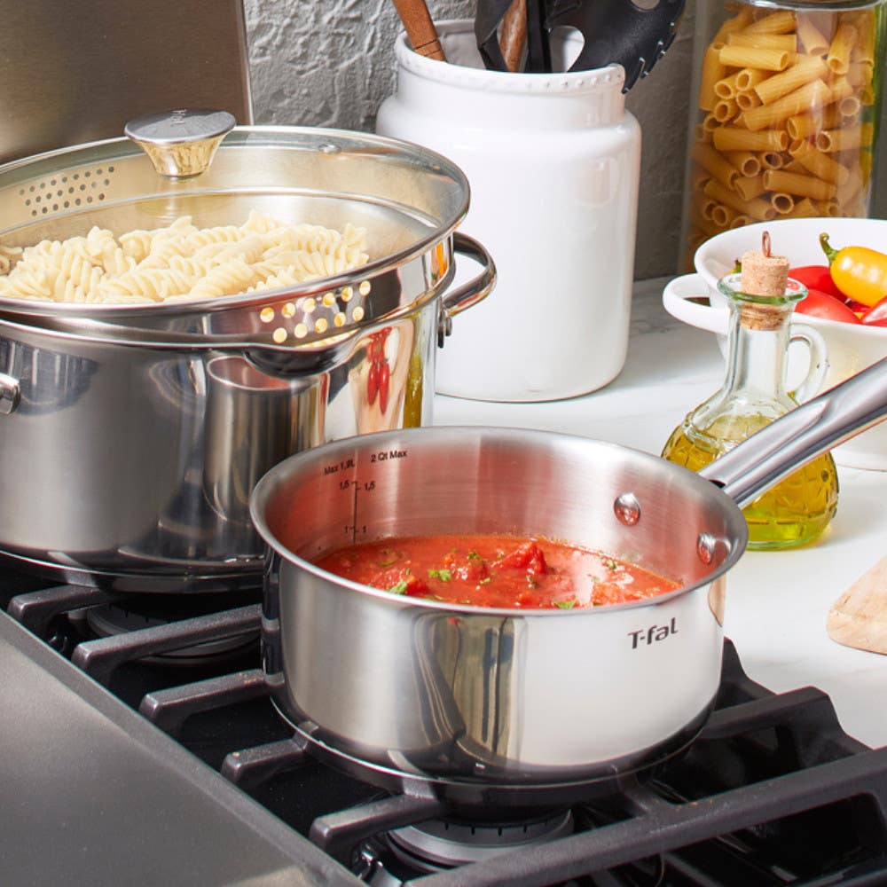 Two stainless steel pots on a stove, one boiling pasta and the other cooking tomato sauce, with cooking utensils and ingredients around them.