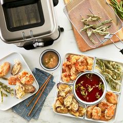 A silver deep fryer with fried tempura vegetables and shrimp arranged on plates, alongside dipping sauces and chopsticks.