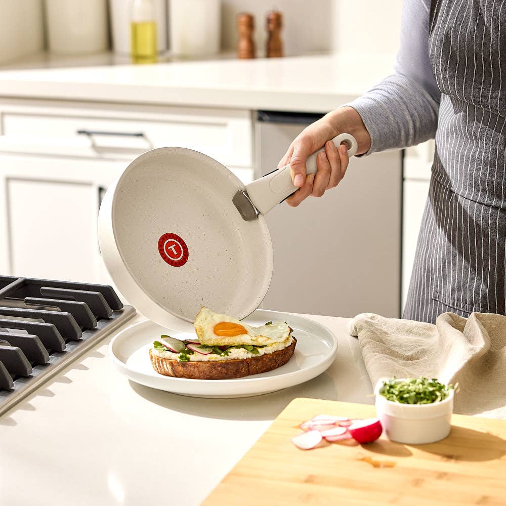 Hand sliding a fried egg from a beige pan with detachable handle onto toast with avocado