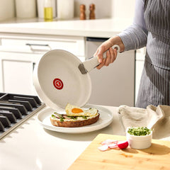 Hand sliding a fried egg from a beige pan with detachable handle onto toast with avocado