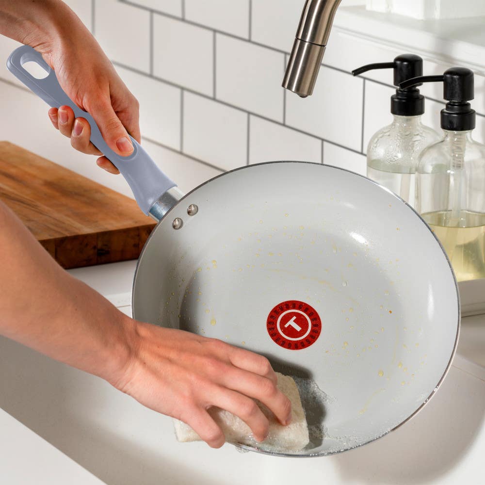 A person handwashing a blue ceramic frying pan with a sponge under a kitchen sink