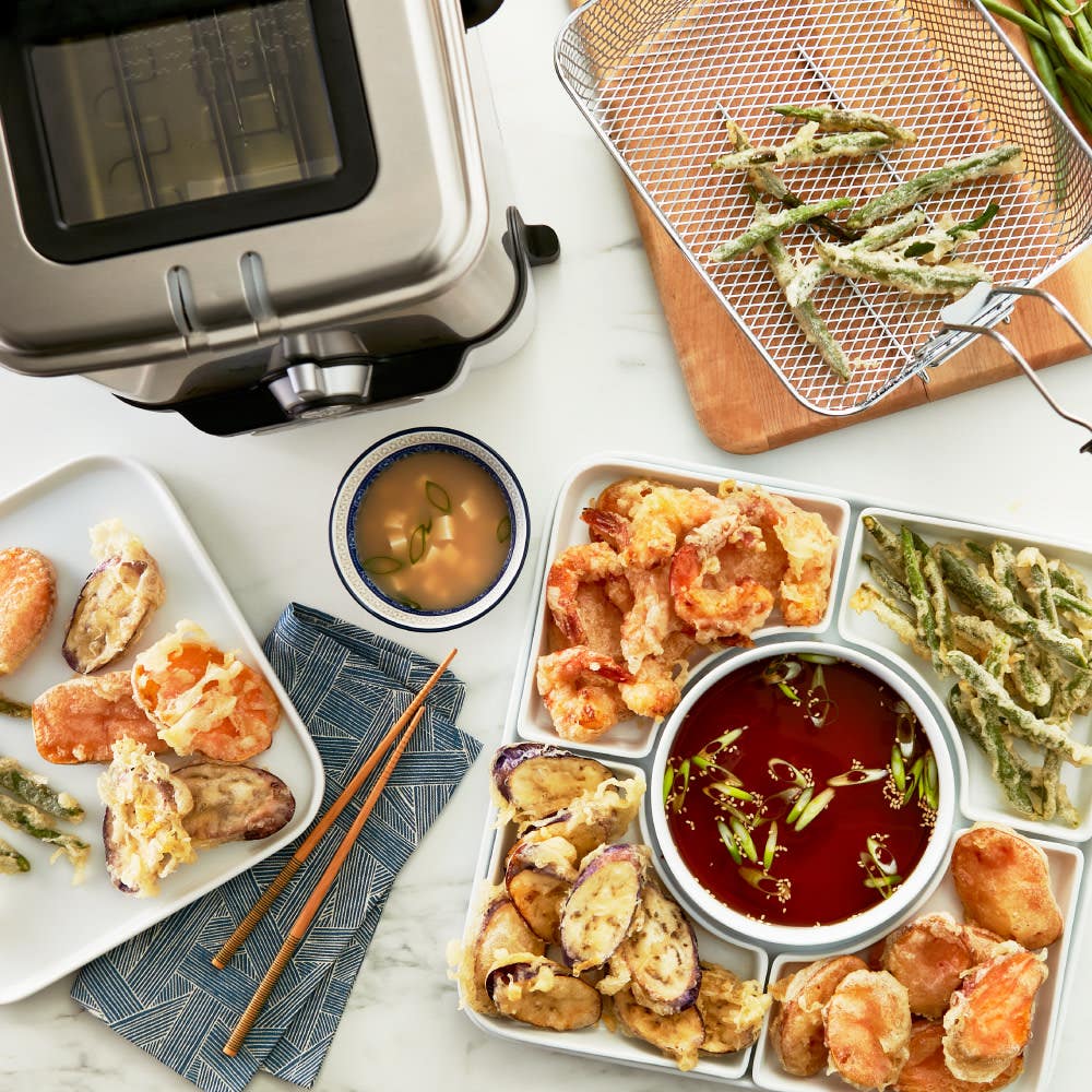 A silver deep fryer with fried tempura vegetables and shrimp arranged on plates, alongside dipping sauces and chopsticks.