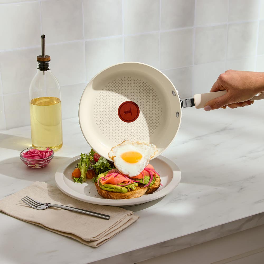 Hand sliding fried egg from a beige nonstick frypan onto a avocado toast on marble countertop