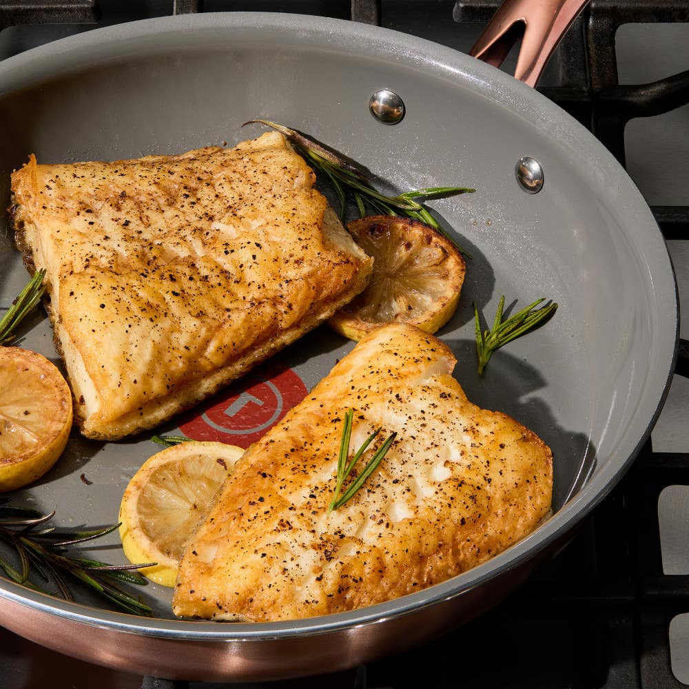 Two seasoned fish fillets cooking in a frying pan with sliced lemons and rosemary sprigs, placed on a gas stove.