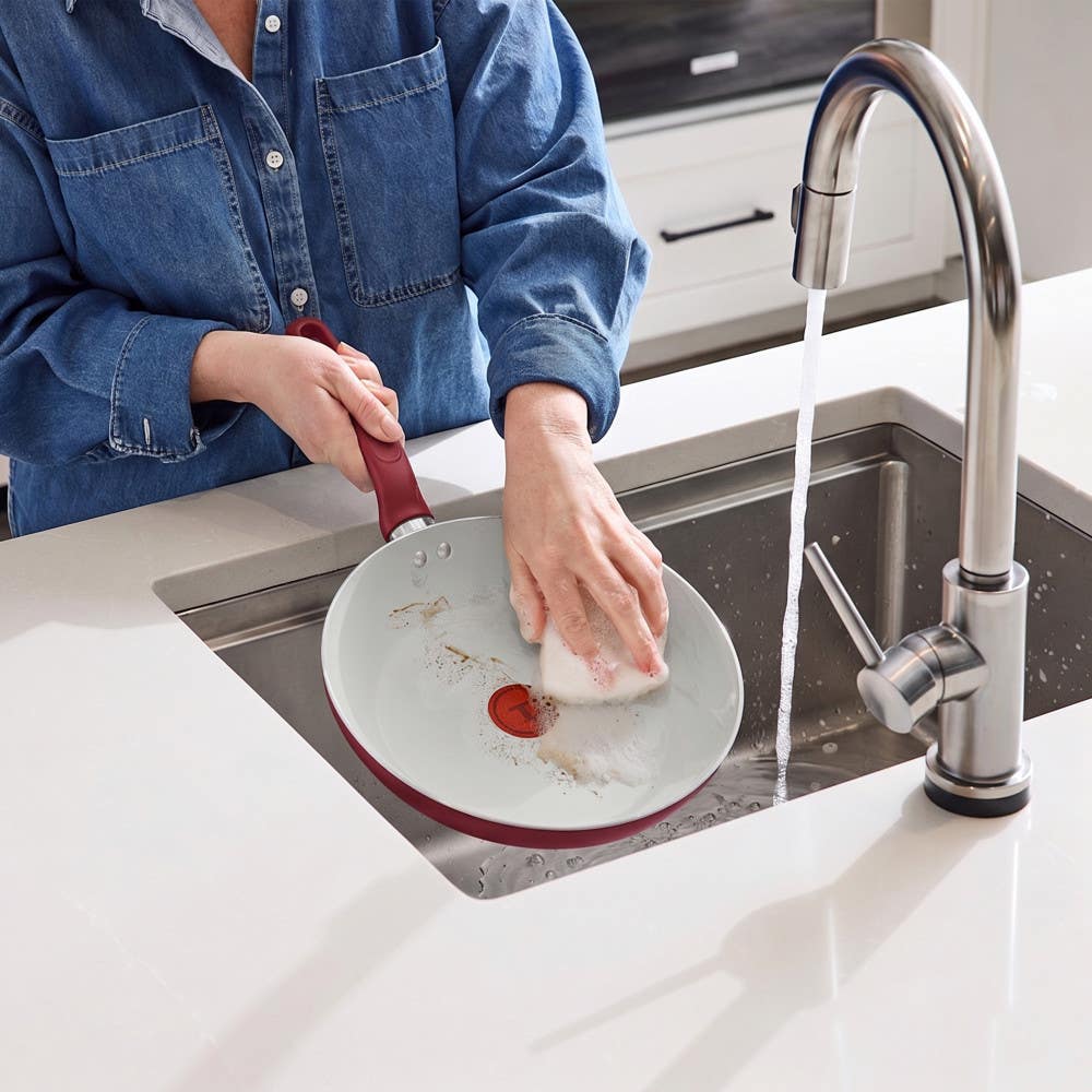 Person washing red frying pan with sponge under running kitchen faucet