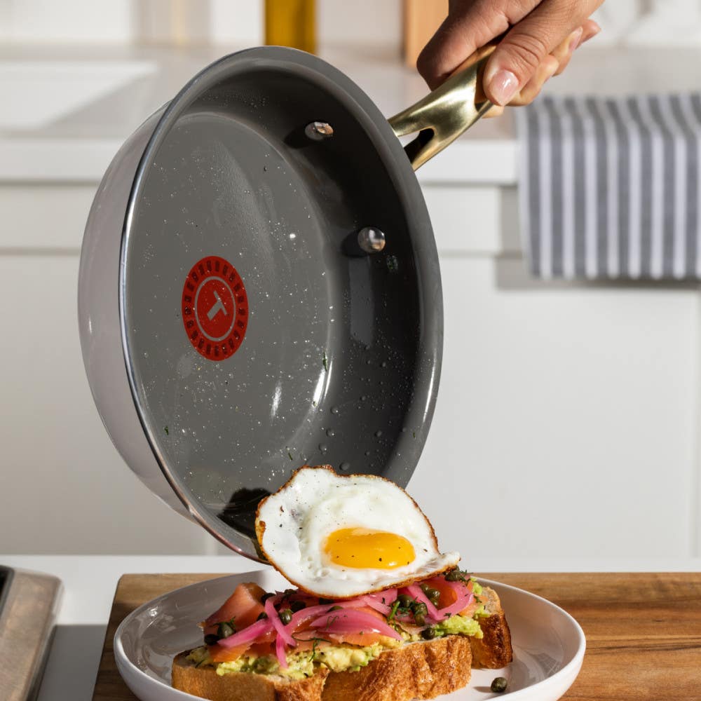 A hand holding a frying pan, tipping a fried egg onto a piece of toast topped with avocado and pink onion slices on a plate.