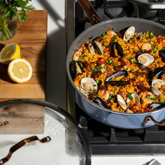 A jumbo cooker on a gas stove filled with seafood paella. Mussels, shrimp, and peas are visible. Sliced lemons sit on a wooden cutting board nearby.