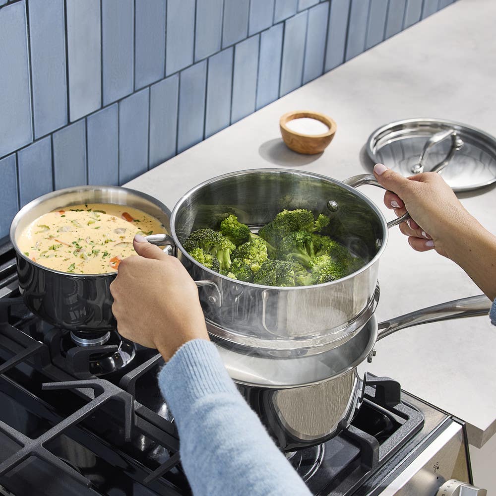 Image of a person steaming broccoli in a stainless steel pot and preparing soup on a gas stove, kitchen setting with blue tile backsplash.