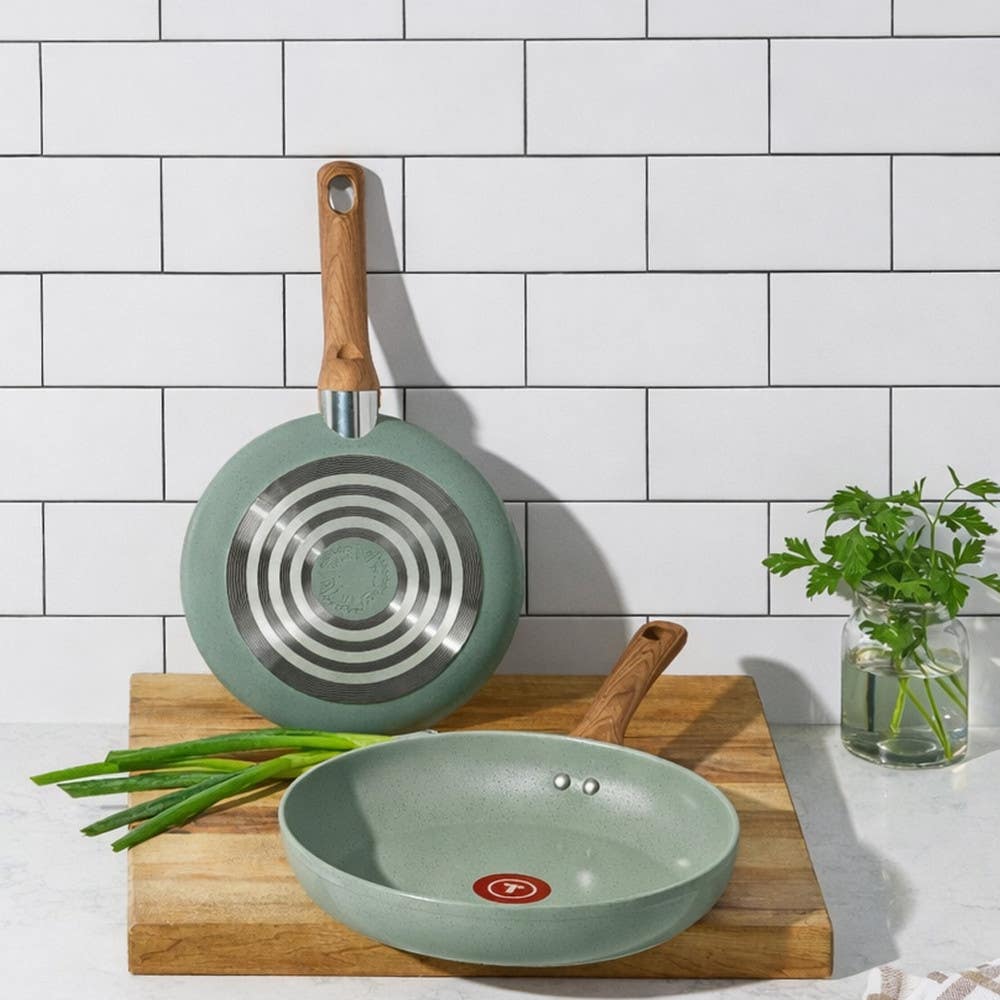 Two green T-fal ceramic frypans, one on board, one upright showing the induction base