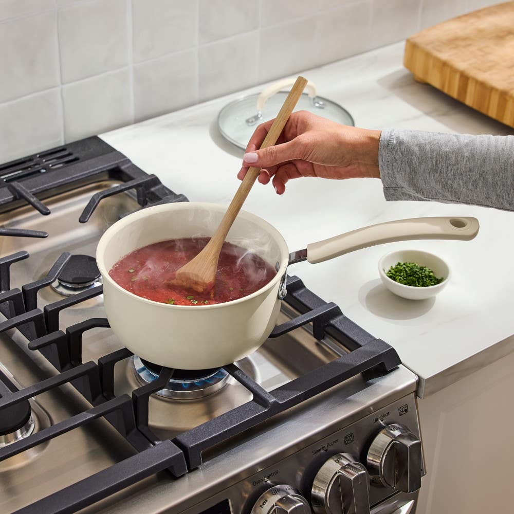 Hand stirring red sauce in beibe nonstick saucepan on lit gas stove with wooden spoon