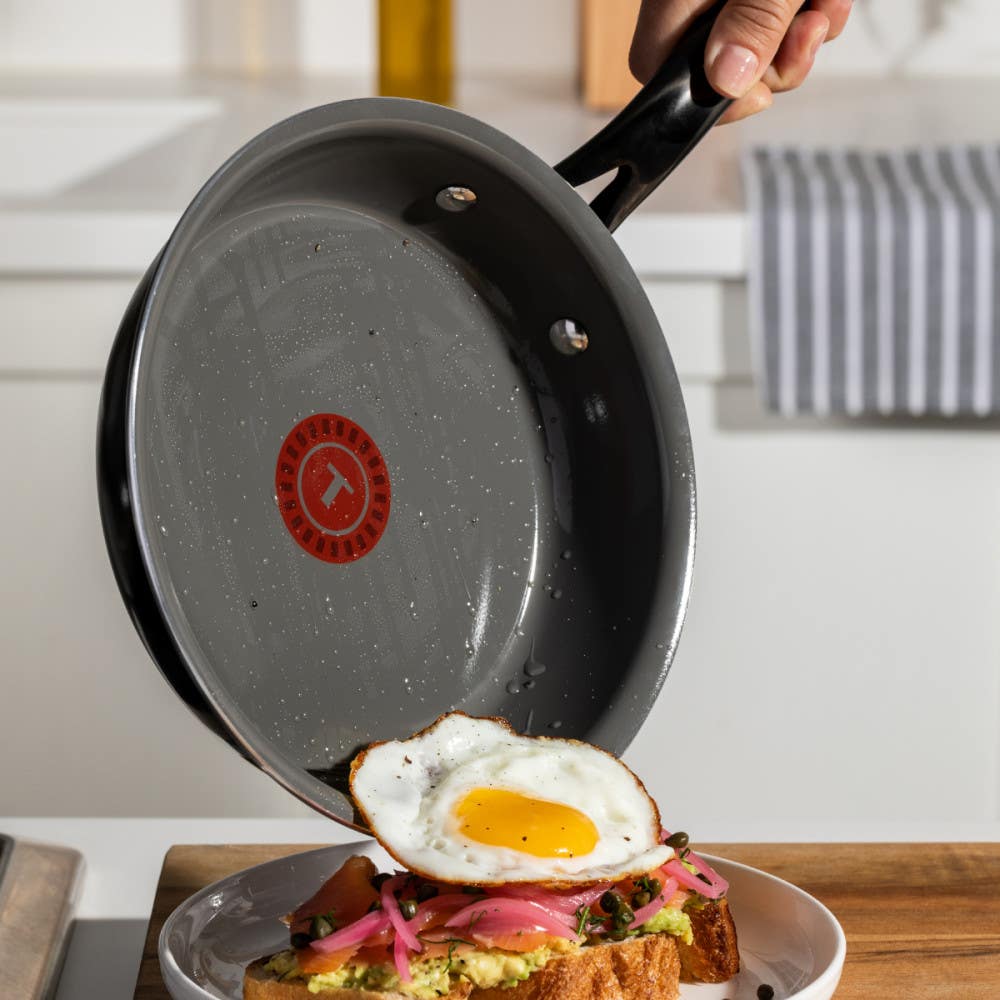 A hand holding a frying pan, tipping a fried egg onto a piece of toast topped with avocado and pink onion slices on a plate.
