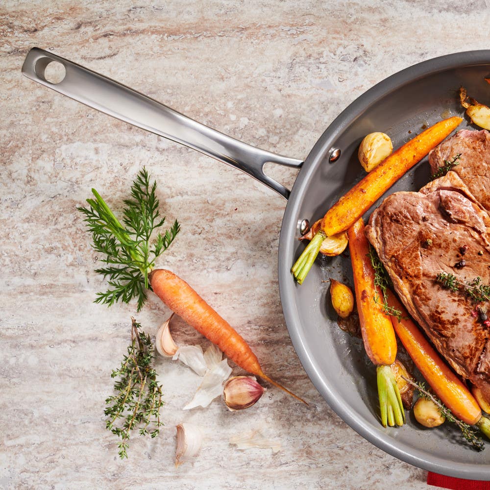 A non-stick frying pan with cooked steak and vegetables, next to fresh ingredients on the counter.