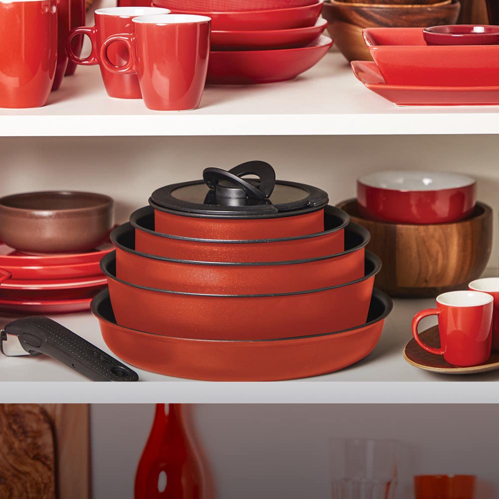 Stacked red cookware among red and white dishware in a cabinet and a detachable handle