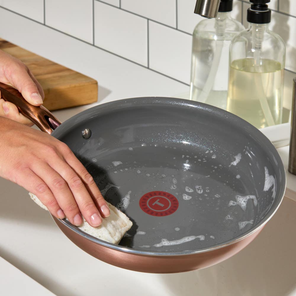 Hand scrubs the interior of a grey pan with a white sponge in a tiled kitchen, with soap dispensers nearby.