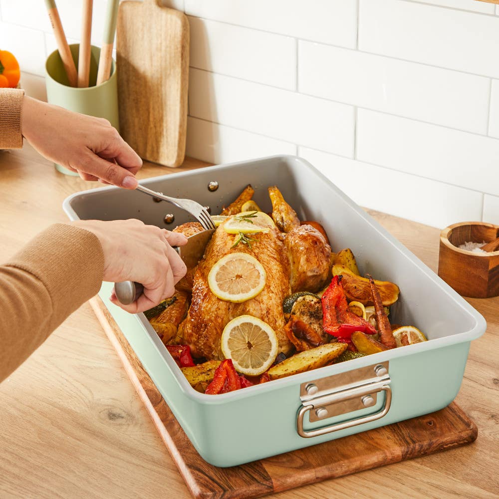Hands carving roasted chicken with vegetables in metal roasting pan.