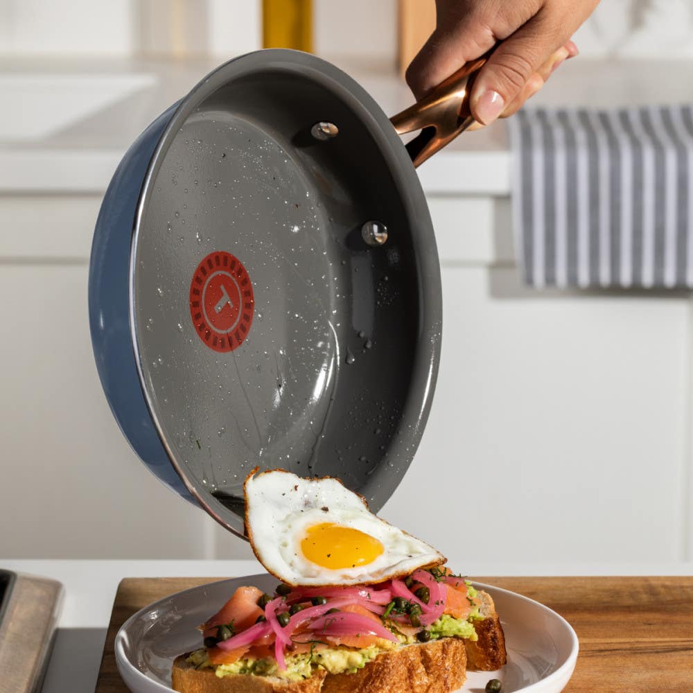 A hand holding a frying pan, tipping a fried egg onto a piece of toast topped with avocado and pink onion slices on a plate.