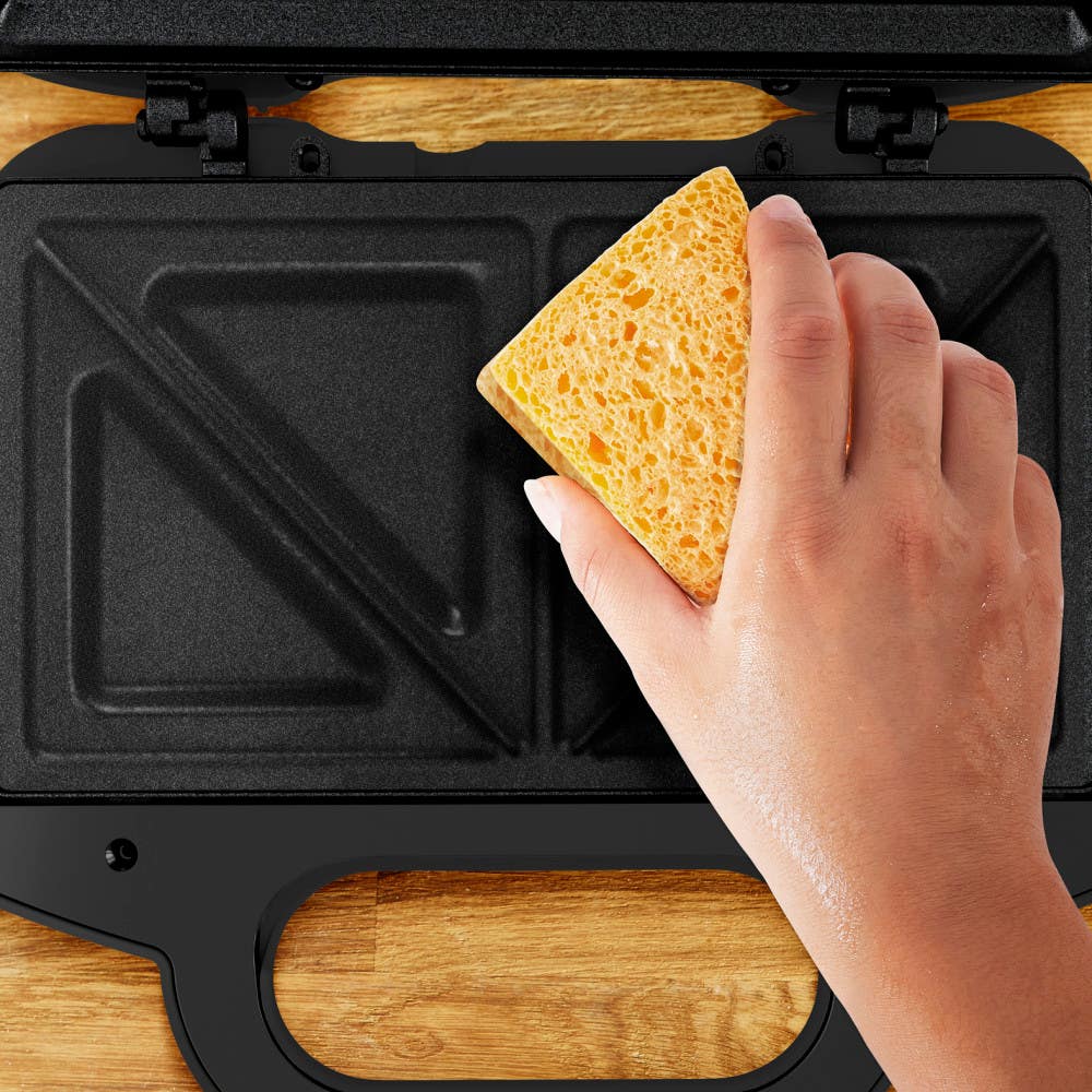 A hand cleaning a sandwich maker's non-stick, triangular-shaped plates with a yellow sponge.