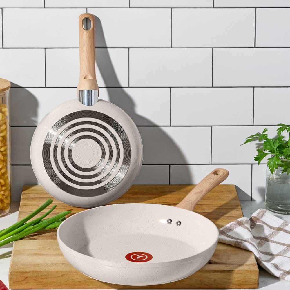 Two beige ceramic frypans with wooden handles on a kitchen counter with ingredients nearby