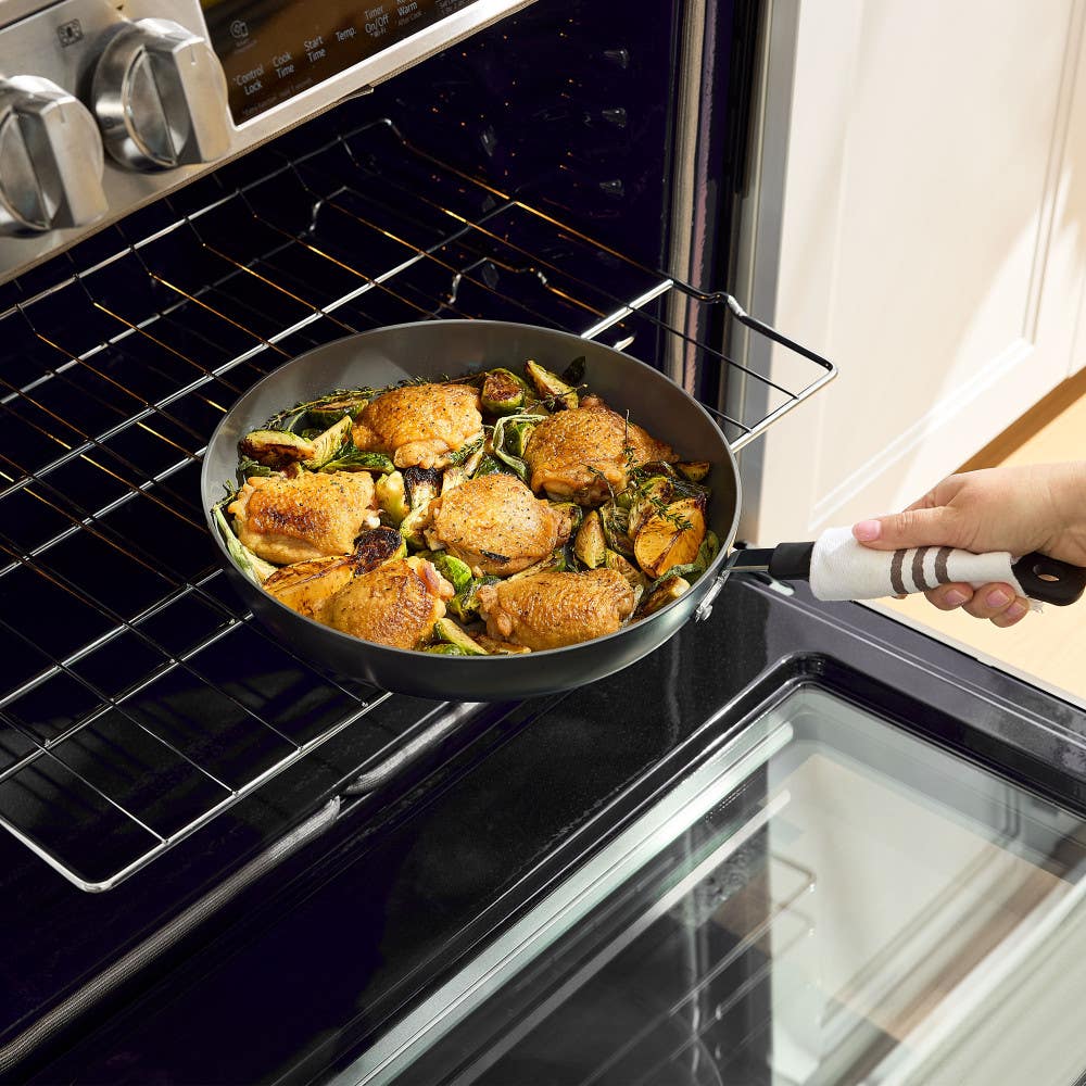 Person placing a ceramic skillet with roasted chicken and vegetables into an oven.