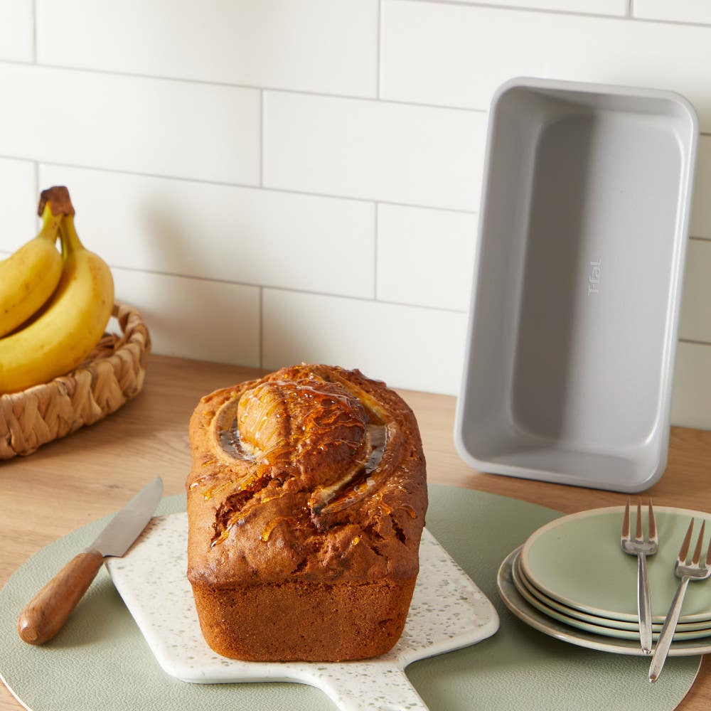 Freshly baked banana bread with a basket of bananas and plates in a kitchen scene.