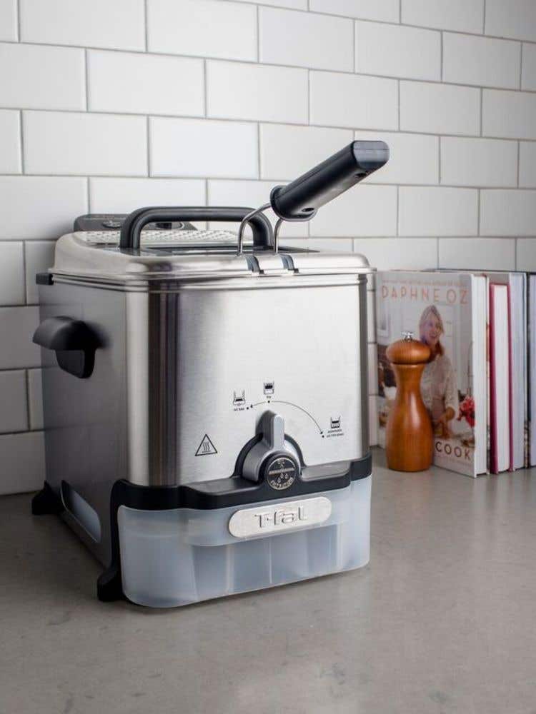 Deep fryer on a countertop, surrounded by cookbooks and a wooden pepper mill. The fryer is stainless steel with a black handle and control panel.