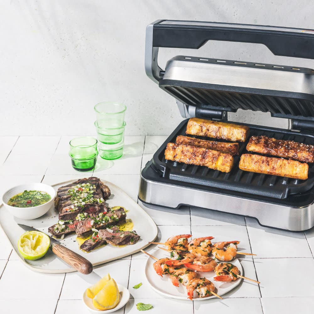Optigrill with skewers and ribs, alongside dishes of grilled meat and seafood on a table.