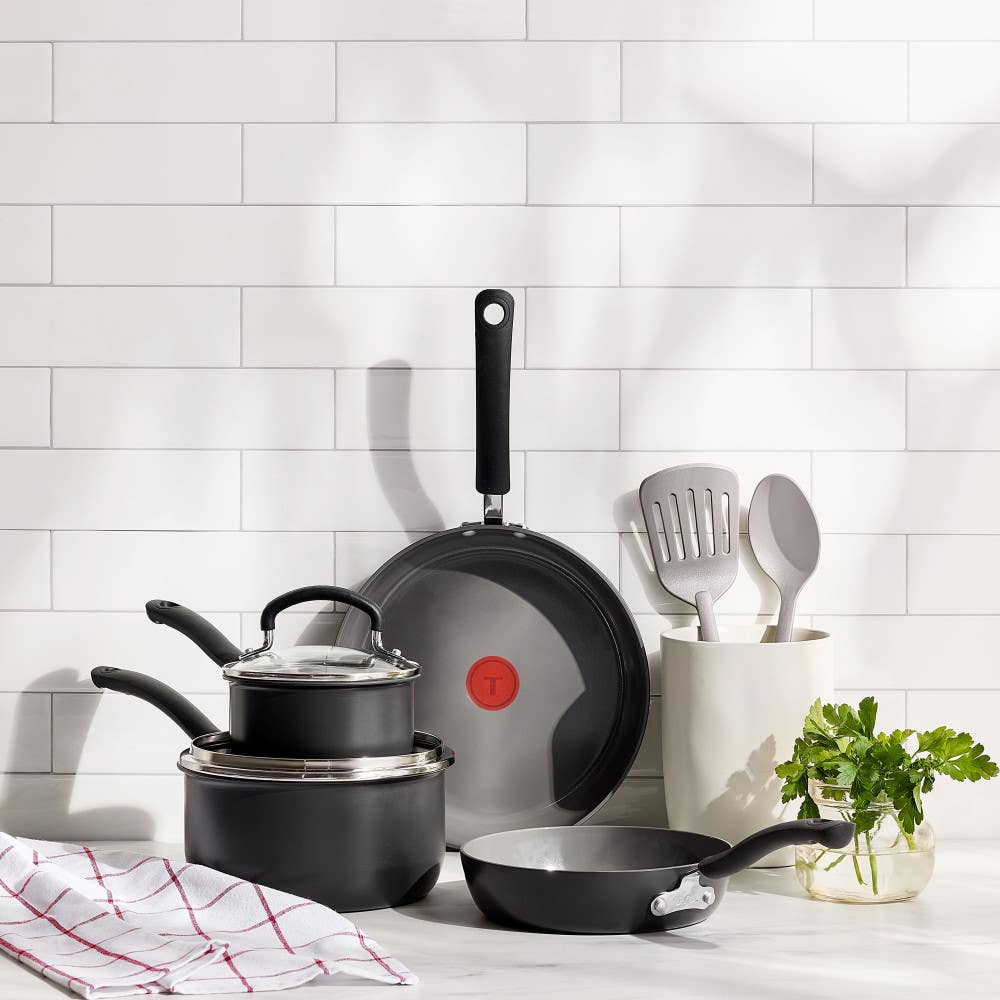 Set of black ceramic cookware and nylon utensils on a kitchen counter with white tile backsplash.