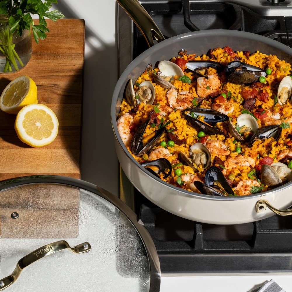 A jumbo cooker on a gas stove filled with seafood paella. Mussels, shrimp, and peas are visible. Sliced lemons sit on a wooden cutting board nearby.