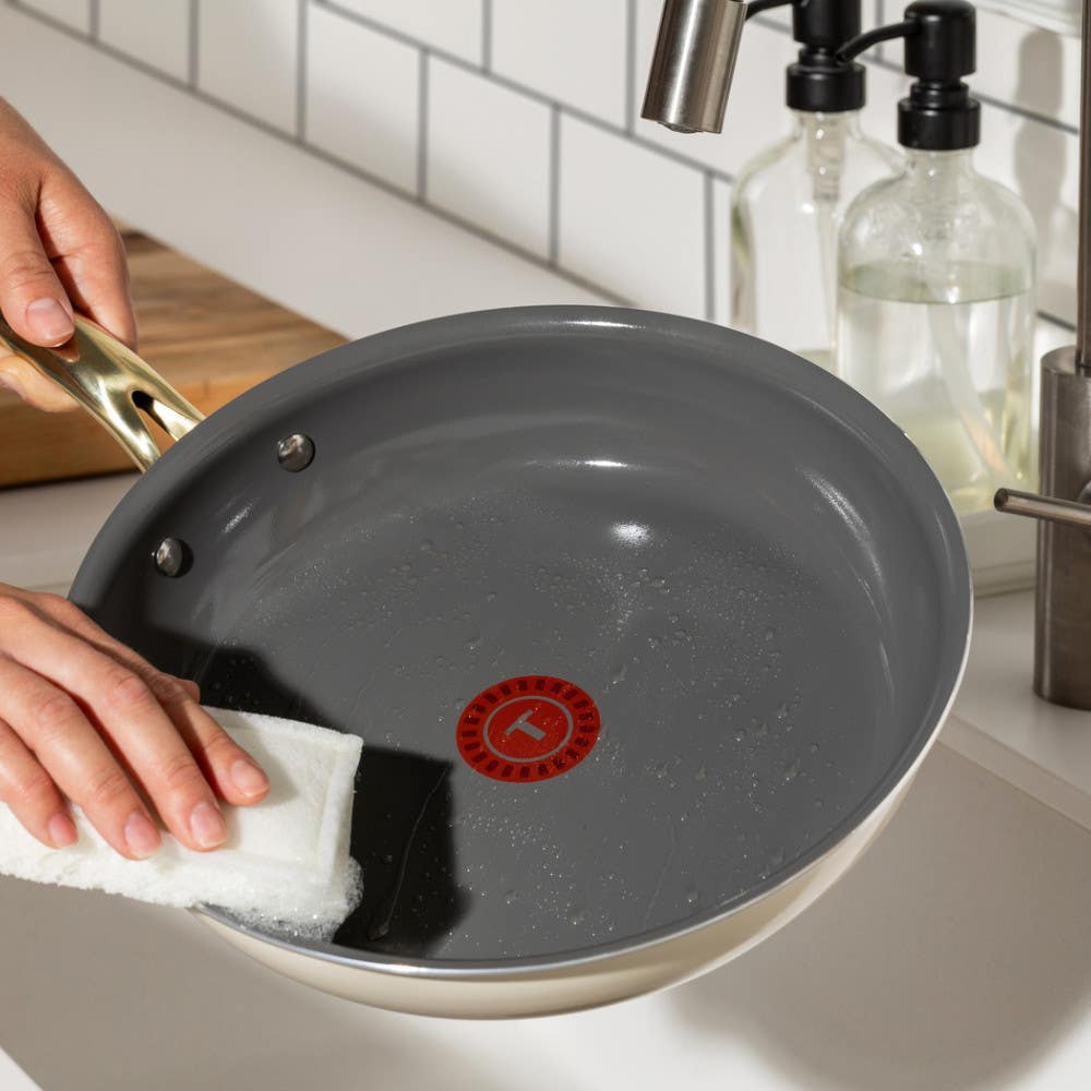 Person cleaning the inside of a pan with a white sponge, showing T-fal's red heat indicator in the center on a kitchen counter.