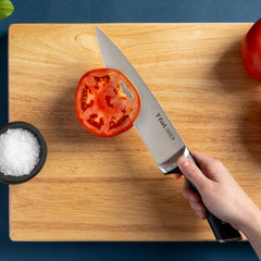 Hand holding a T-fal knife slicing a tomato on a wooden cutting board.