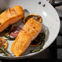 Two salmon fillets cooking in a ceramic frypan with lemon slices and herbs on a stove