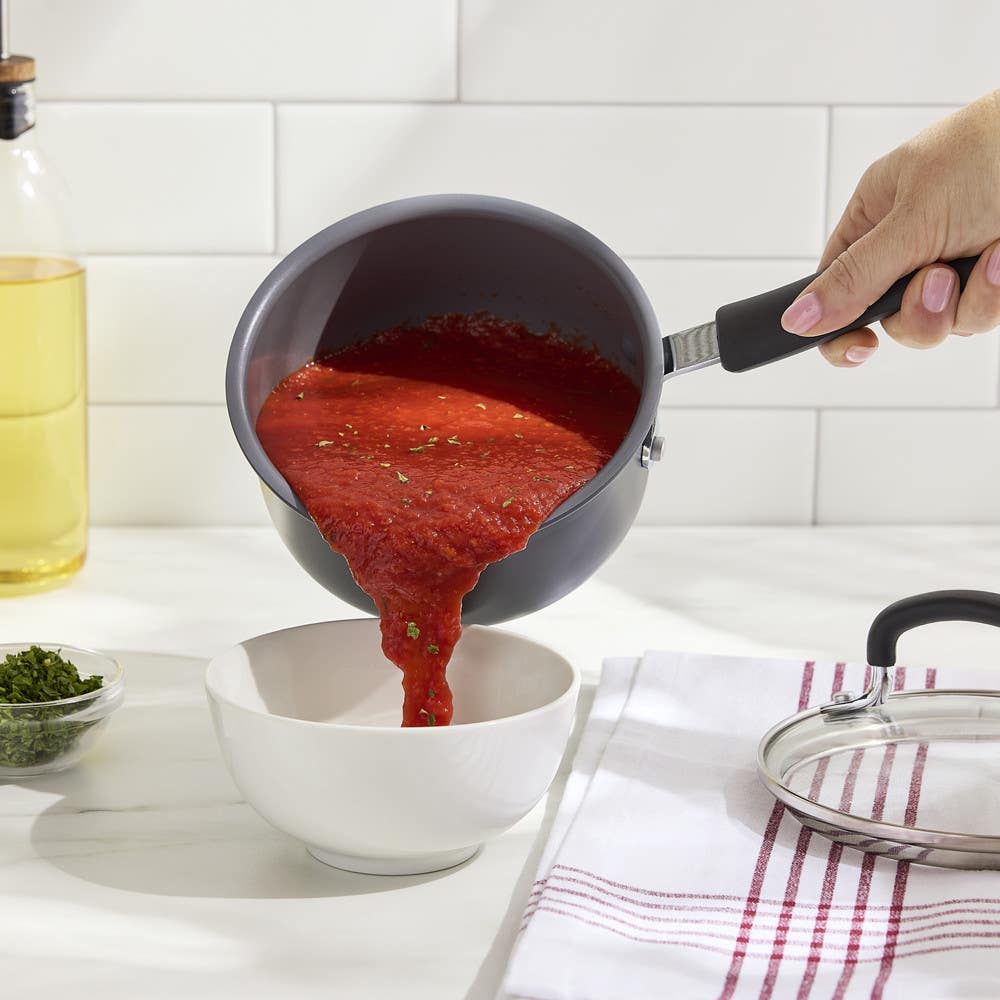Hand pouring tomato sauce from nonstick saucepan into a white bowl on a kitchen counter.