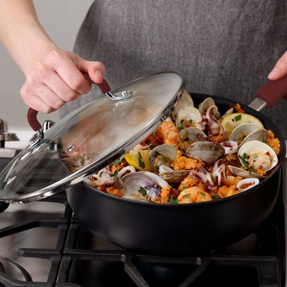 Person lifting lid off a jumbo cooker with red handle filled with seafood stewing on a stove.