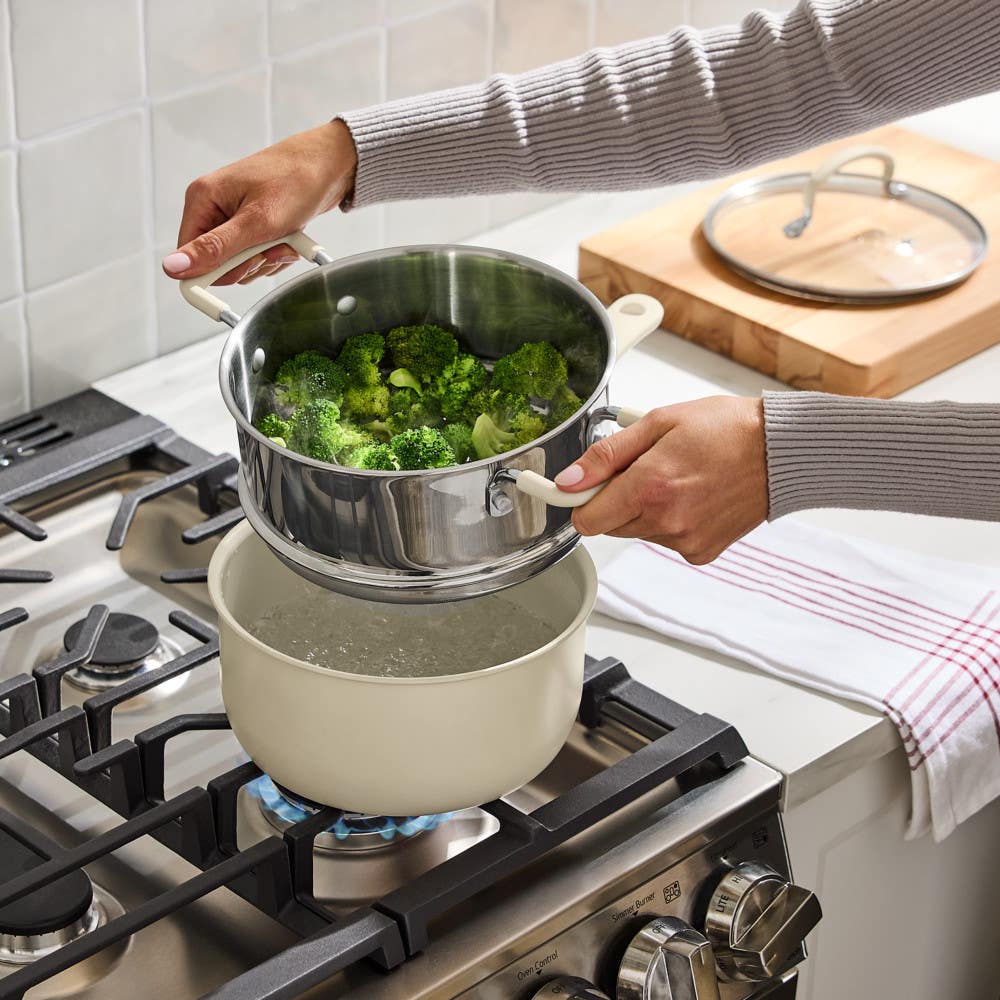 Hands lifting steamer with broccoli from beige nonstick stockpot on lit gas stove in modern kitchen