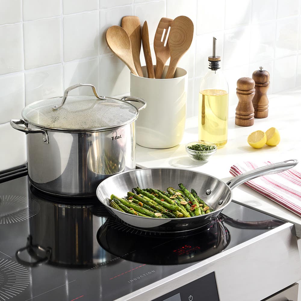 Stainless steel cookware set on a modern stovetop with asparagus in the frying pan.