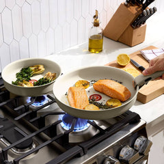 Two beige frying pans with detachable handles on a gas stove, cooking fish and vegetables
