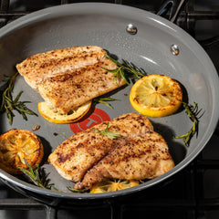Two seasoned fish fillets cooking in a frying pan with sliced lemons and rosemary sprigs, placed on a gas stove.
