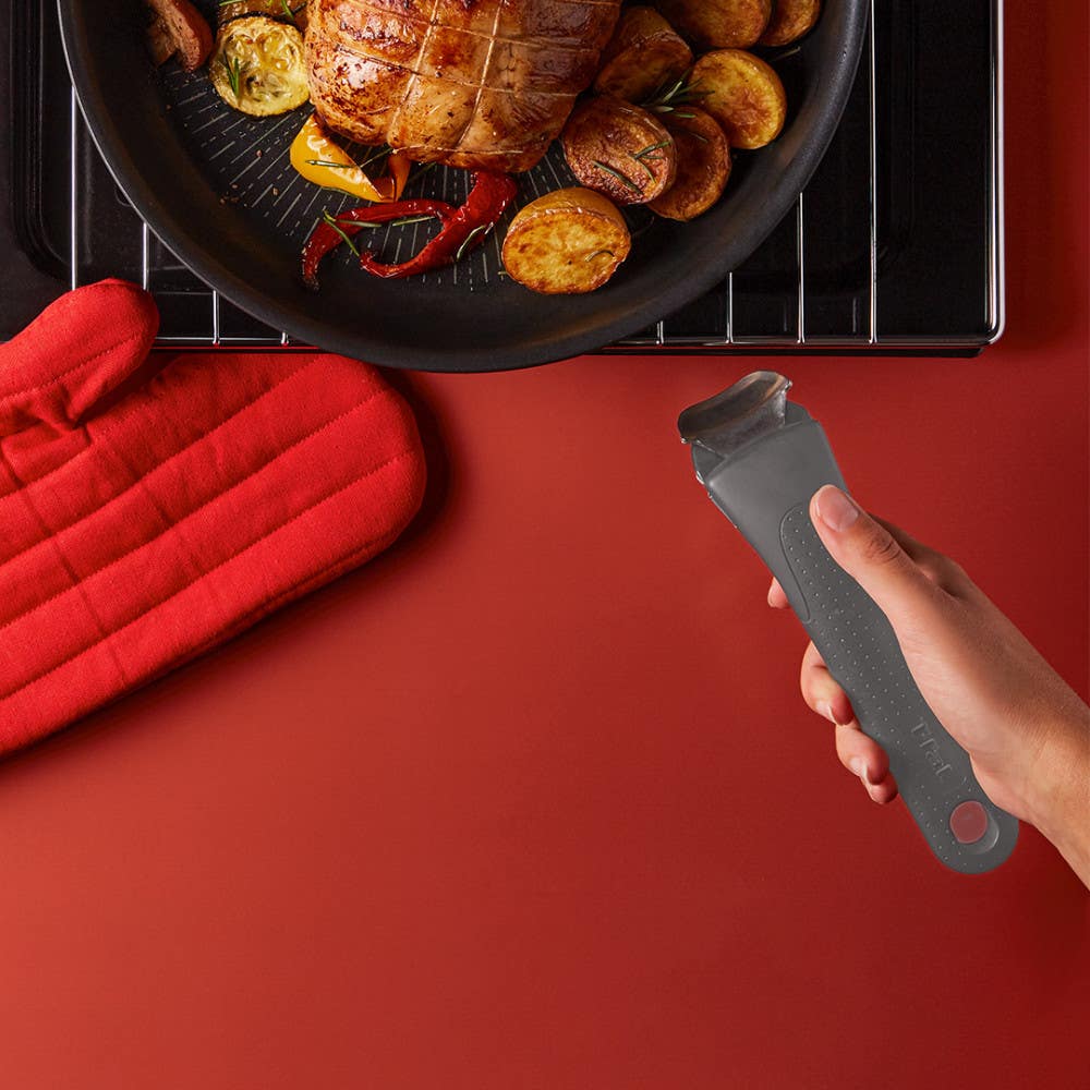Black pan with roasted food, a red oven mitt, and a hand holding a grey detachable handle