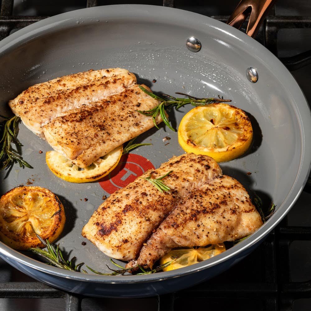 Two seasoned fish fillets cooking in a frying pan with sliced lemons and rosemary sprigs, placed on a gas stove.